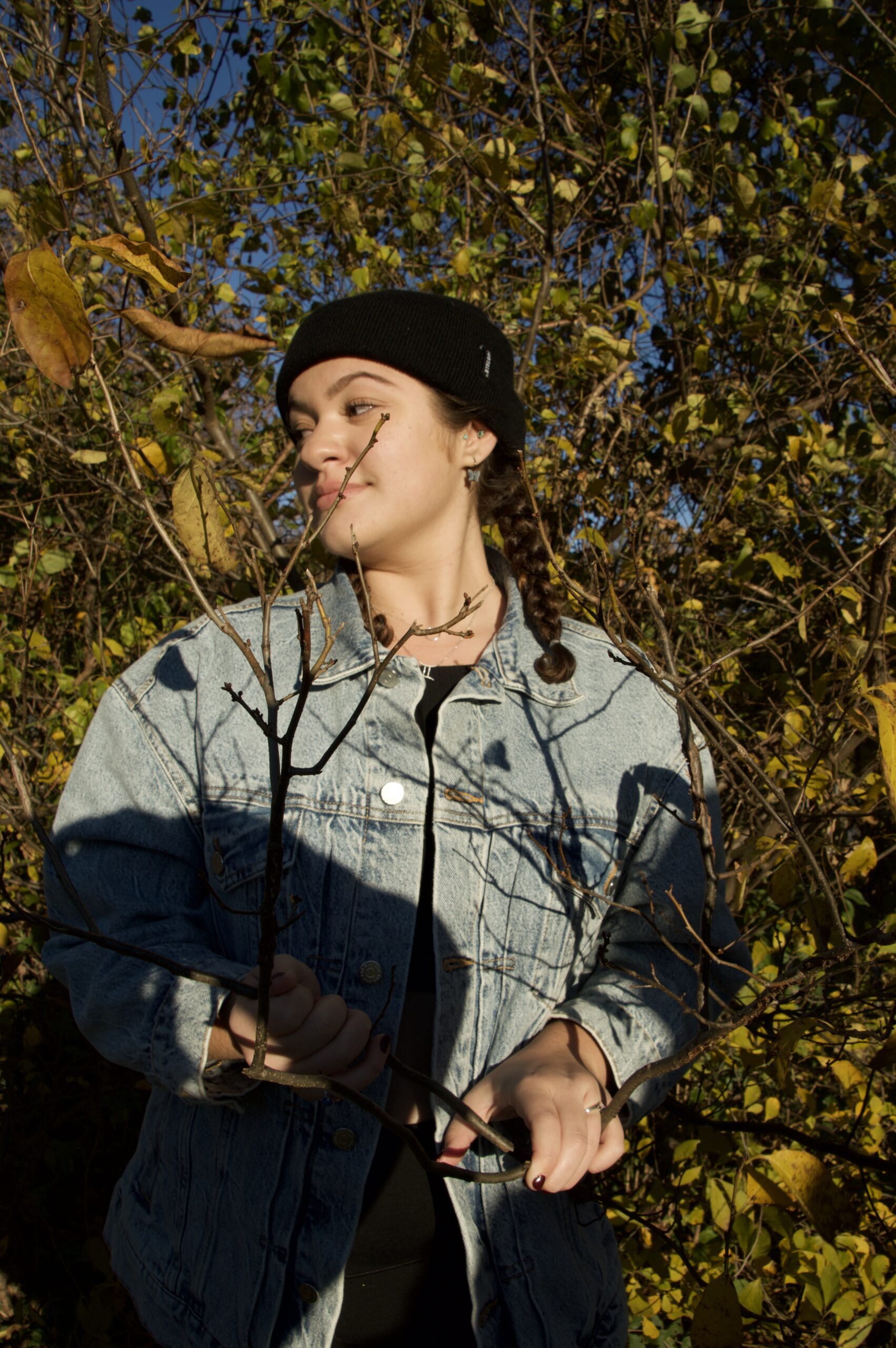 Girl in a denim jacket and a black beanie looks to the left as her hands wrap around tree branches. She is surrounded by green and yellow leaves and trees